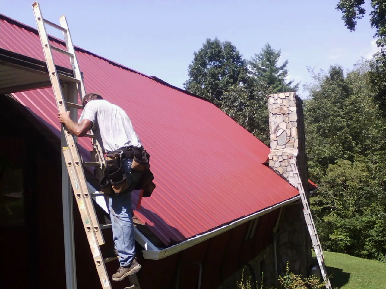 Roofer on a ladder beside a red metal roof and stone chimney — Soffit & Fascia Repair by Roofing Brothers in Still Pond